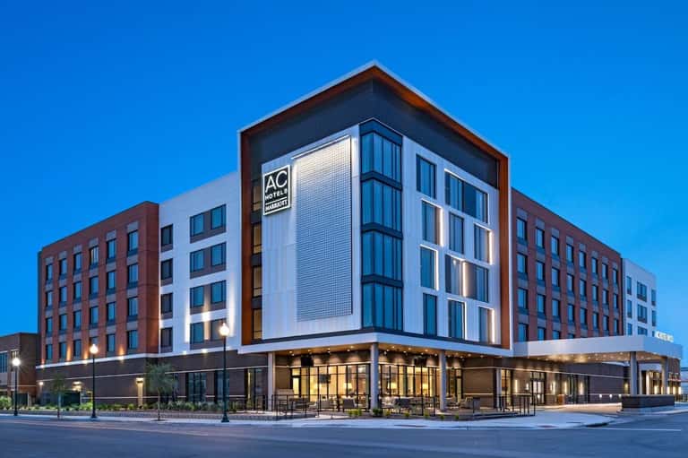 Contemporary AC Hotel building exterior with red brick, white panels, and illuminated ground-floor entrance at dusk