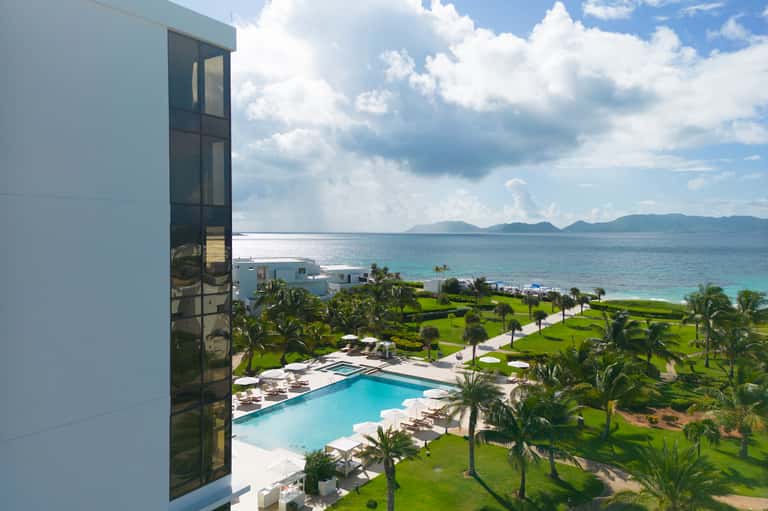Beachfront resort view from hotel window showing ocean, pool, tropical gardens, palm trees, and distant mountains