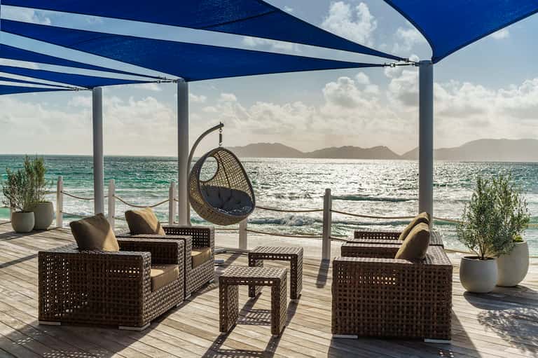 Beachfront lounge area with woven furniture under blue sail canopy overlooking ocean and mountains