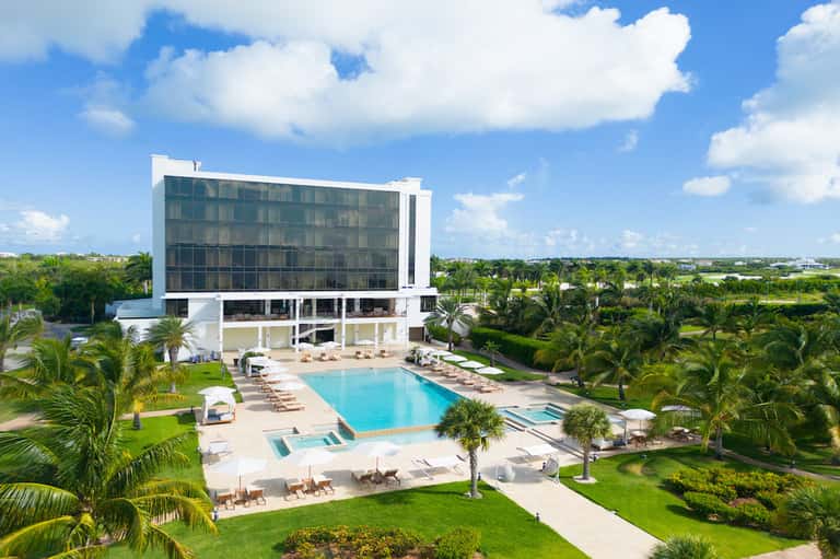 Modern resort exterior with turquoise pool, palm trees, and manicured grounds under blue sky