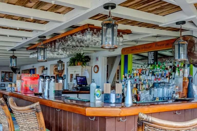 Rustic wooden bar counter with hanging glassware, colorful bottles, and nautical-themed décor