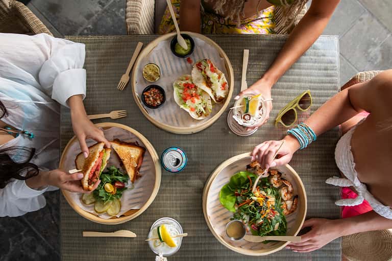 Overhead view of people dining outdoors with tacos, sandwiches, and salads on wooden plates