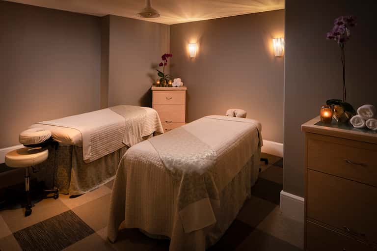 Spa treatment room with two massage tables draped in cream linens and soft ambient lighting