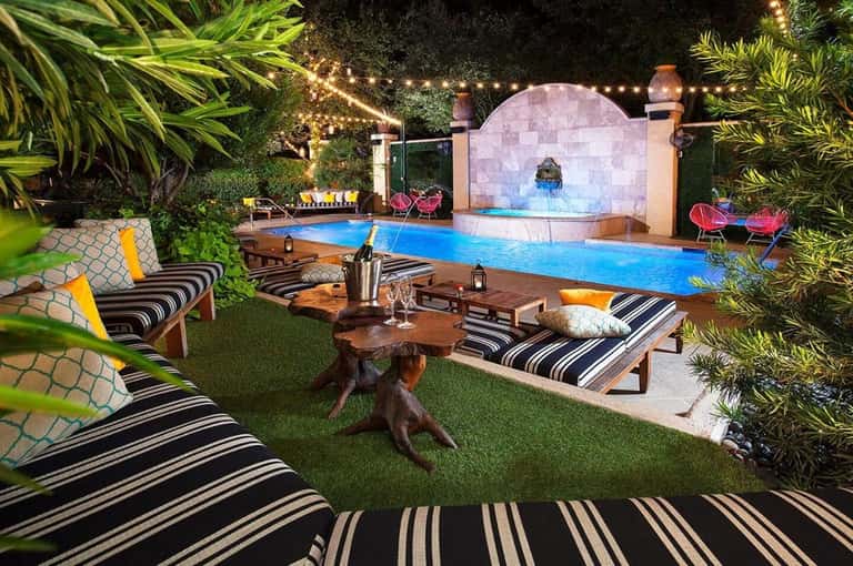 Nighttime resort pool area with striped loungers, tropical plants, illuminated water features, and fire pit