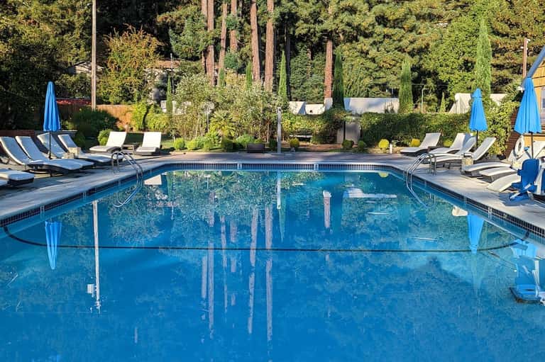 Luxury pool surrounded by tall trees, blue umbrellas, and lounge seating area