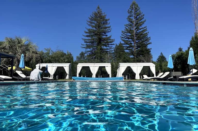 Resort outdoor pool with crystal-clear water, white cabanas, blue umbrellas, and tall pine trees in background