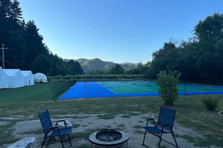 Outdoor sports facility with blue tennis courts, white tent structures, fire pit, and forested mountain landscape