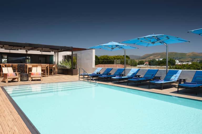 Resort rooftop pool with turquoise water, blue lounge chairs, blue umbrellas, and mountain views in background