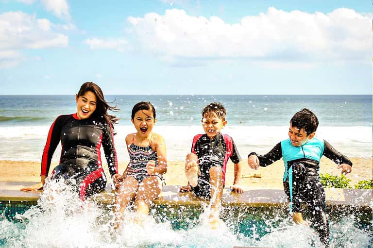 Four children in colorful swimwear running through shallow water at beach with ocean backdrop
