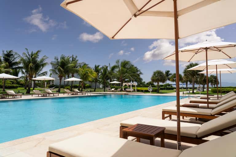 Resort pool area with lounge chairs, umbrellas, palm trees, and manicured grounds