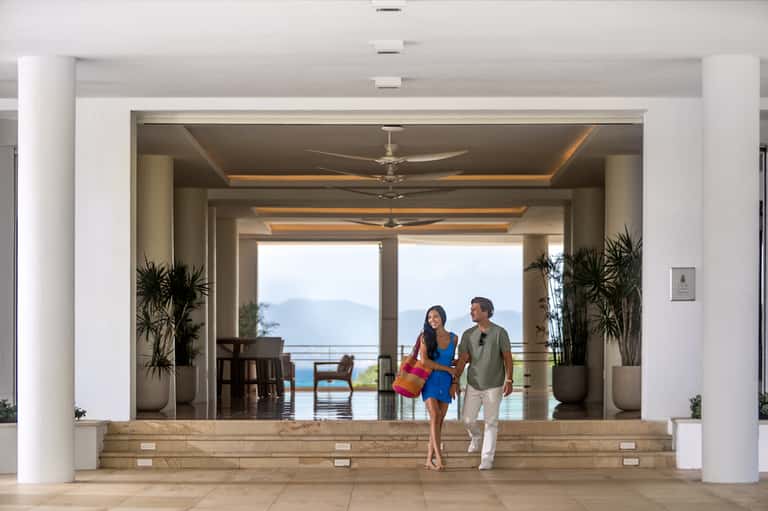 Bright modern lobby with white columns, potted plants, ceiling fans, and two people walking toward an ocean view terrace