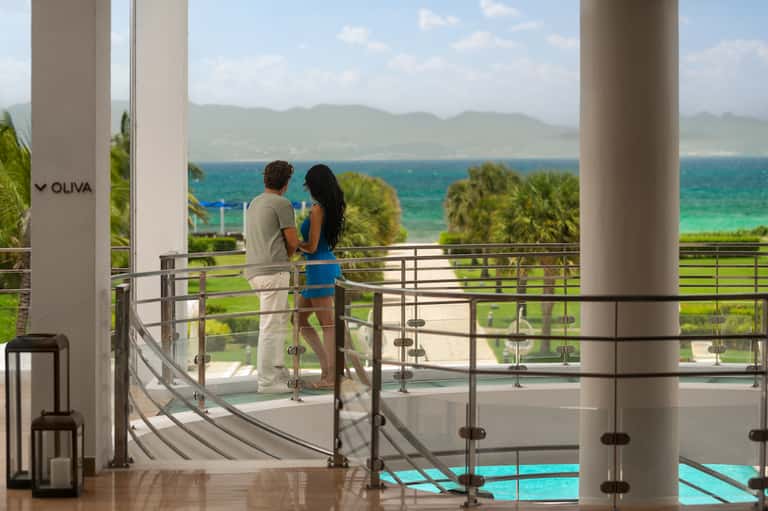 Couple standing on terrace with metal railings overlooking ocean, mountains, and palm trees in coastal setting.