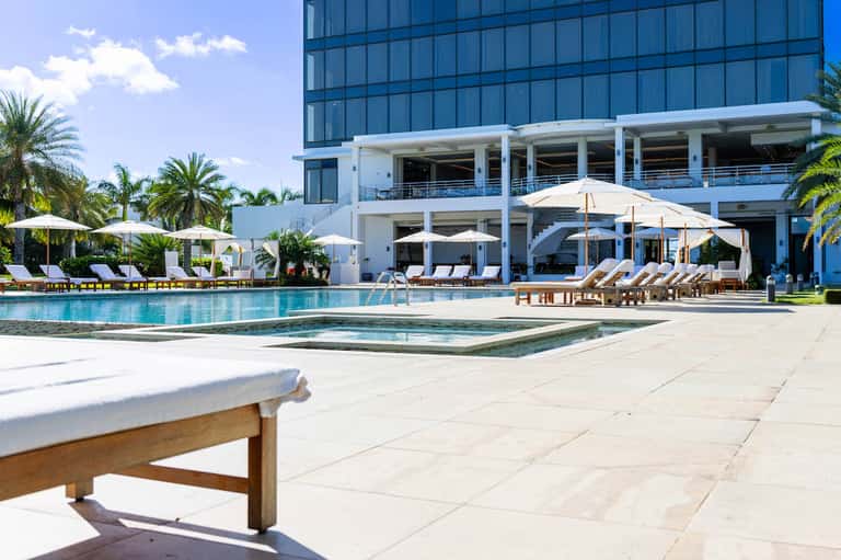 Luxury resort pool deck with white umbrellas, lounge chairs, and modern multi-story hotel building with palm trees