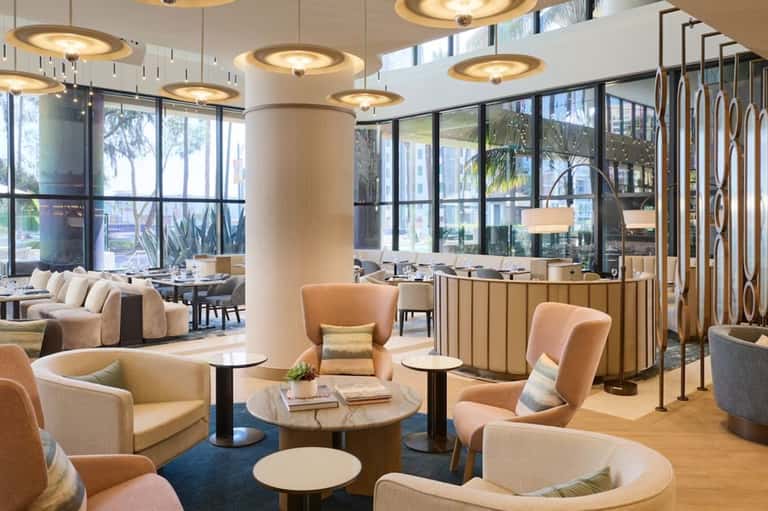 Modern hotel lobby with warm lighting, comfortable seating areas, floor-to-ceiling windows overlooking palm trees and street