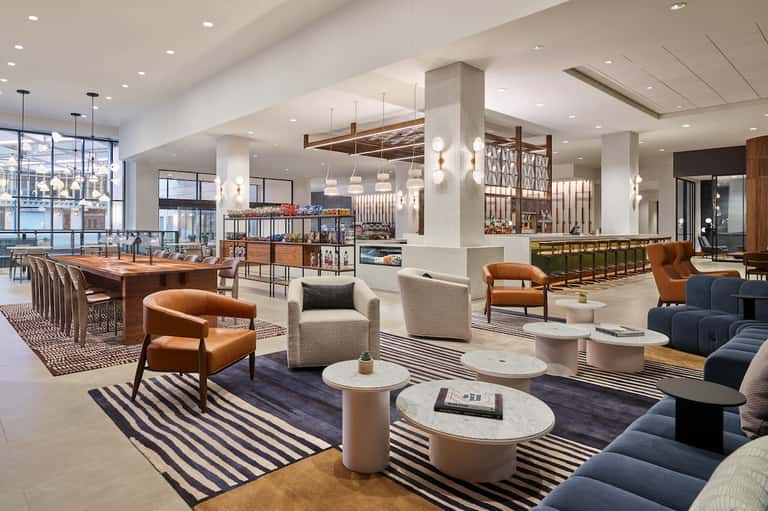 Modern hotel lobby with wood and marble finishes, orange leather chairs, pendant lights, and striped rugs