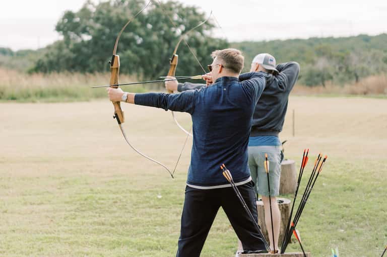 Man practicing archery outdoors with bow and arrows, wearing blue shirt and cap near wooded backdrop