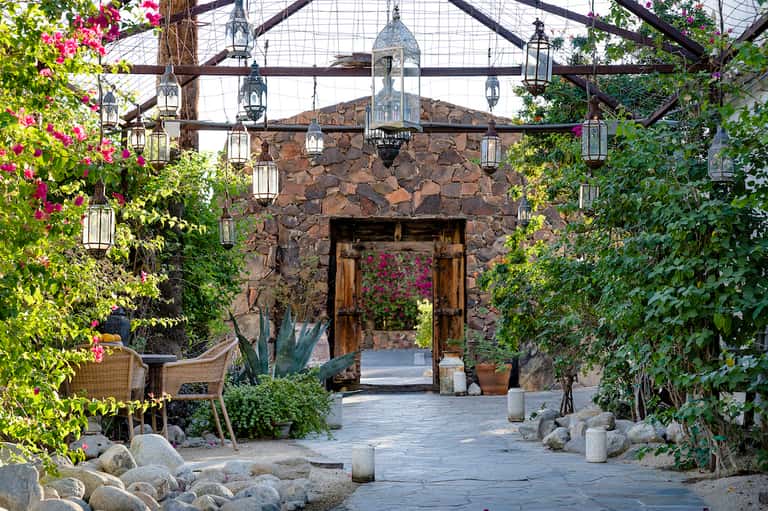 Desert courtyard with stone archway, wooden doors, climbing flowers, hanging lanterns, and decorative boulders
