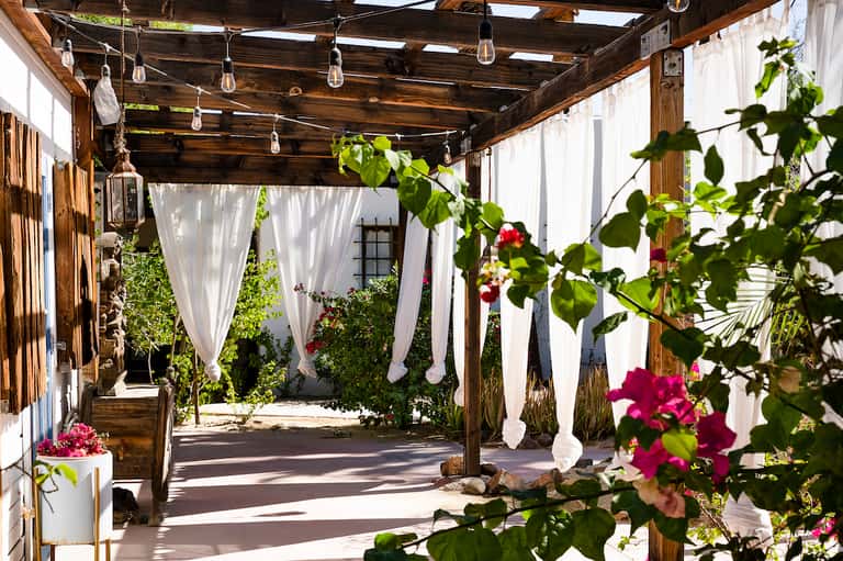 Wooden pergola with white sheer curtains, hanging lights, and climbing magenta flowers in garden setting