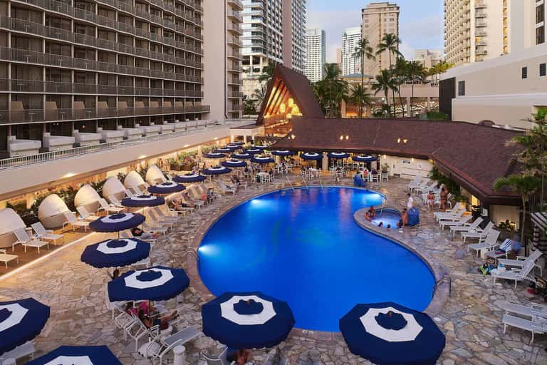 Illuminated resort pool at dusk with blue umbrellas, lounge seating, and high-rise hotel buildings