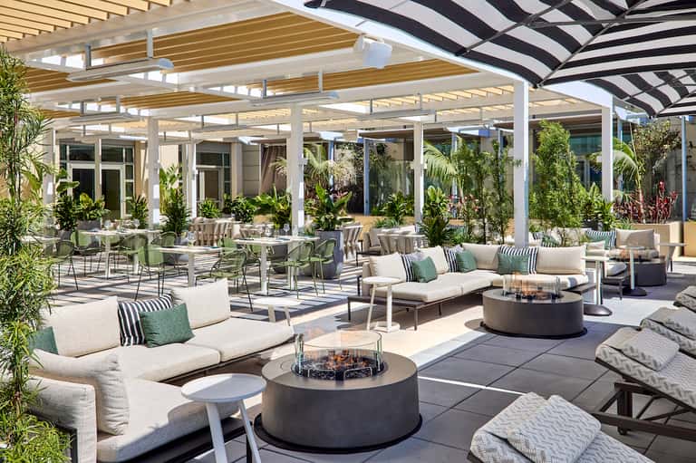 Expansive outdoor patio with lounge seating, fire pit, white pergola with adjustable louvers, striped umbrellas, and tropical landscaping