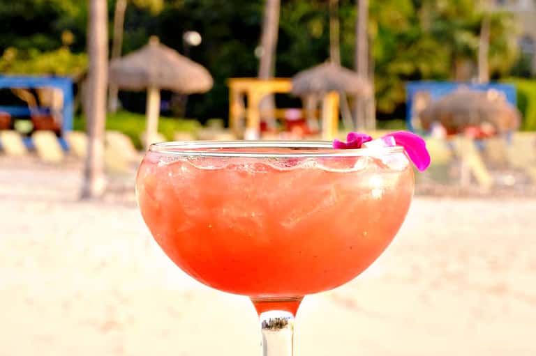 Coral-colored cocktail with ice and pink flower garnish on beach with umbrellas and loungers