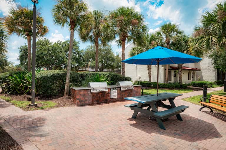 Outdoor patio with picnic table, blue umbrella, grill station, and palm trees
