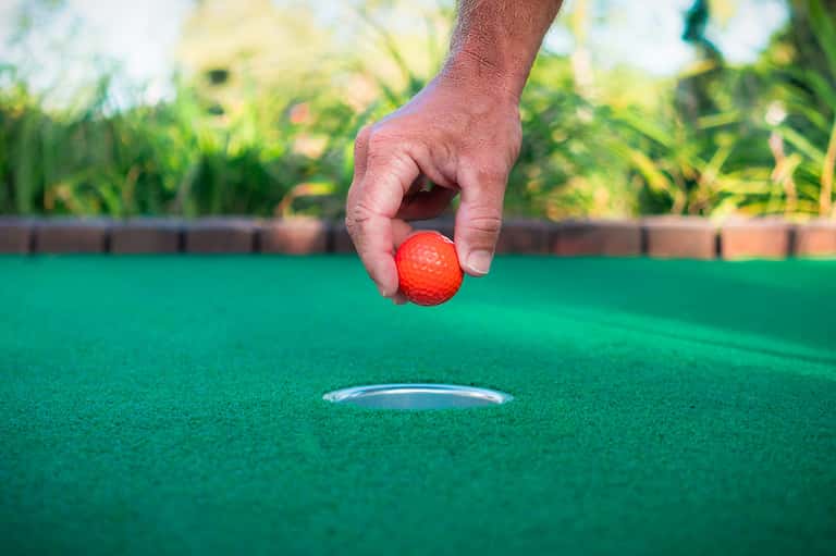Hand holding red golf ball above hole on putting green with blurred garden background