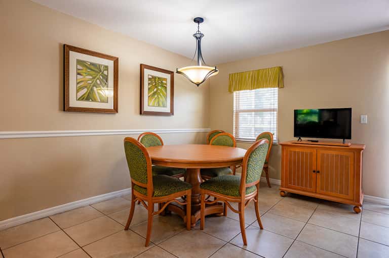Casual dining room with round wood table, green chairs, TV, and tropical artwork