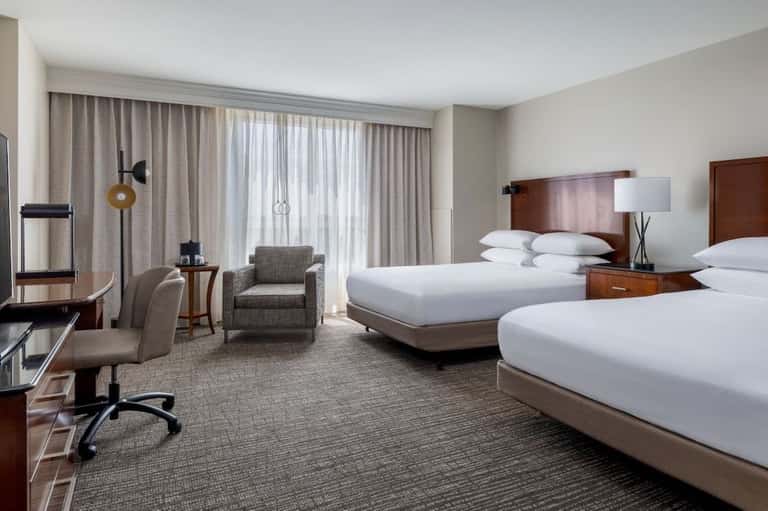 Hotel guest room with two beds, work desk, armchair, and balcony access