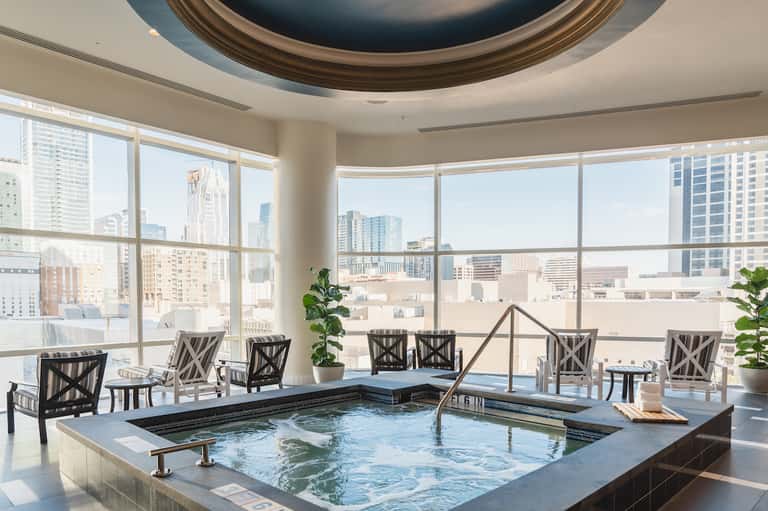 Modern indoor hot tub with city skyline views through floor-to-ceiling windows and lounge seating