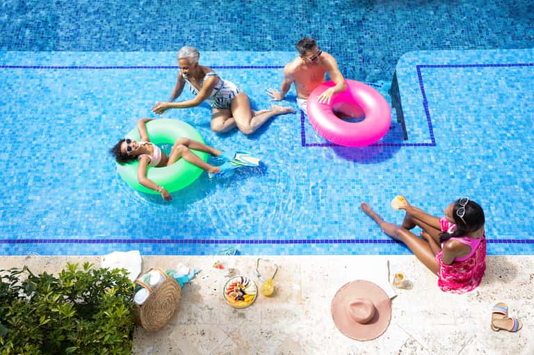 Family in swimming pool with colorful inflatable floats on sunny day with poolside snacks and palm trees