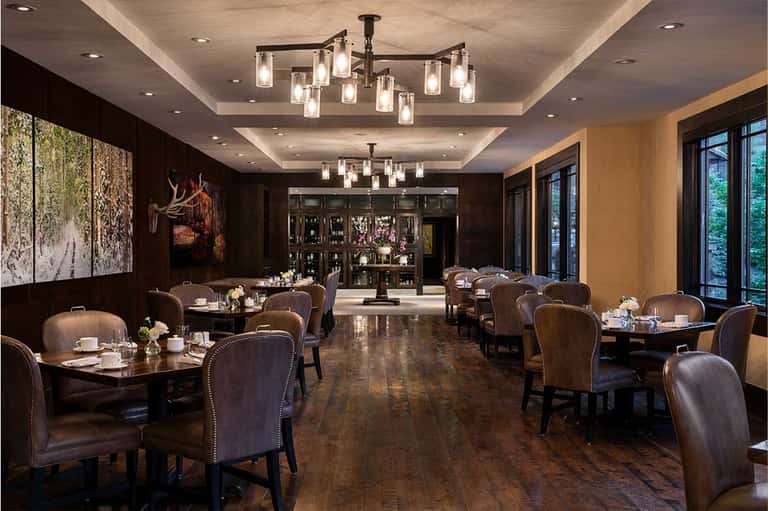 Upscale dining room with dark wood paneling, leather chairs, round tables, chandeliers, and bar area