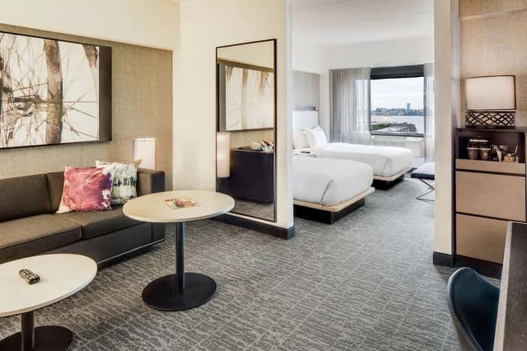 Modern hotel suite with living area, bedroom, and city waterfront view through floor-to-ceiling windows