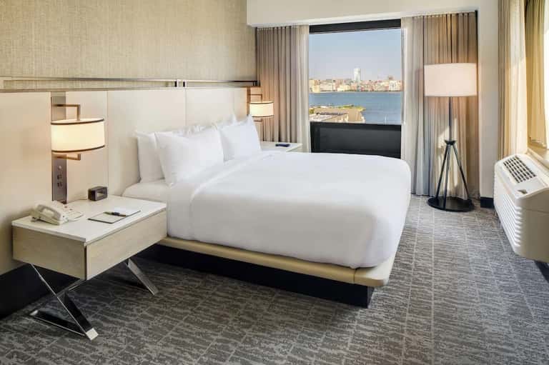 Modern hotel bedroom with queen bed, waterfront city views, floor-to-ceiling windows, and contemporary furnishings