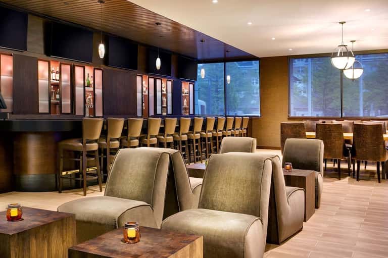 Upscale lounge bar with dark wood cabinetry, tan seating, pendant lighting, and large windows