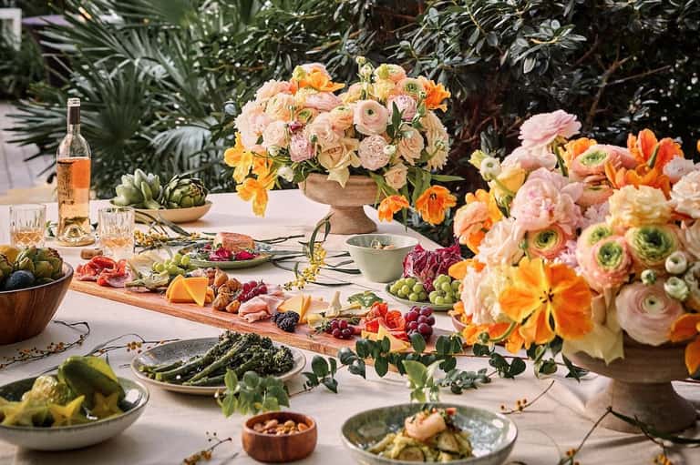 Outdoor dining table with colorful flowers, wine, fresh produce, and Mediterranean cuisine