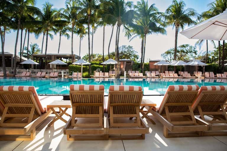 Resort pool deck with wooden lounge chairs, striped orange and white cushions, white umbrellas, and palm trees