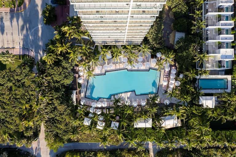 Aerial view of turquoise pool surrounded by palm trees, lounge chairs, and buildings with pergola shade structures