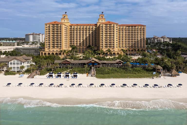 Beachfront resort with twin clock towers, sand beach, and rows of blue umbrellas along shoreline