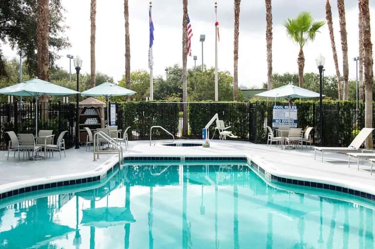 Outdoor pool surrounded by palm trees, lounge chairs, umbrellas, and white fencing with flags