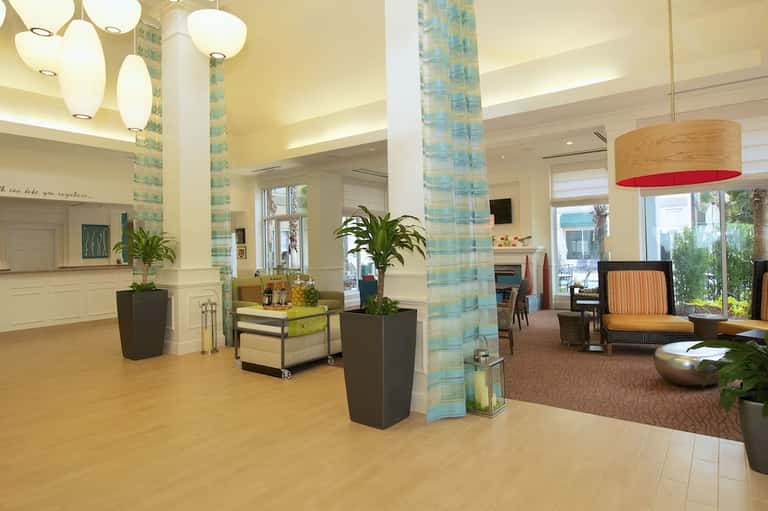 Bright hotel lobby with pendant lights, white columns, plants, and lounge seating areas