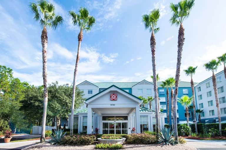 Hotel entrance with white architectural features, palm trees, and blue residential buildings in background