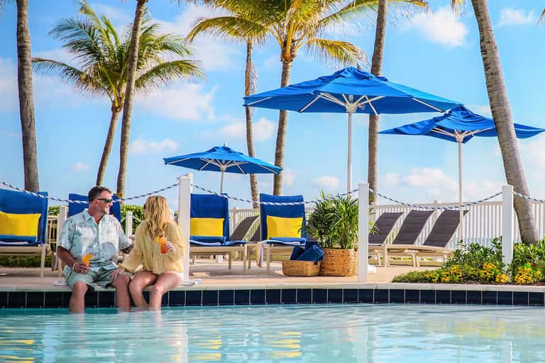 Couple sitting poolside under blue umbrellas with palm trees and resort in background