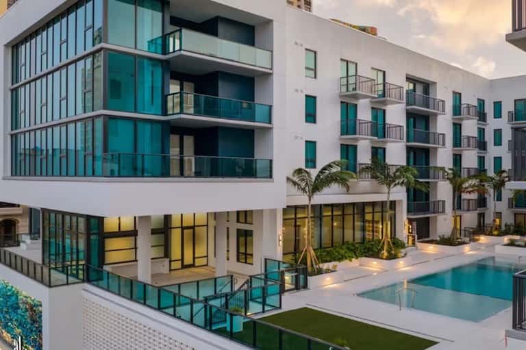 Modern waterfront residential building with white facade, turquoise balconies, pool, and palm trees