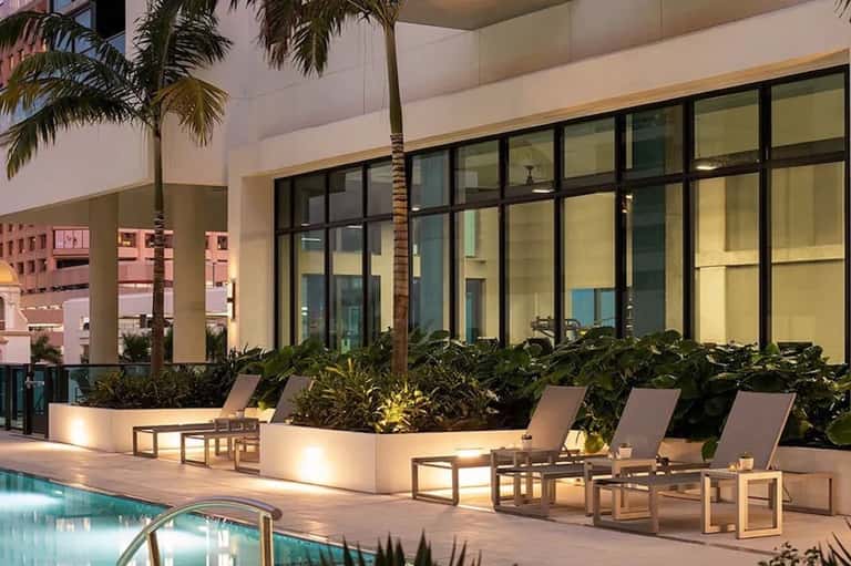 Modern poolside terrace with lounge chairs, illuminated planters, tall windows, and palm trees at dusk