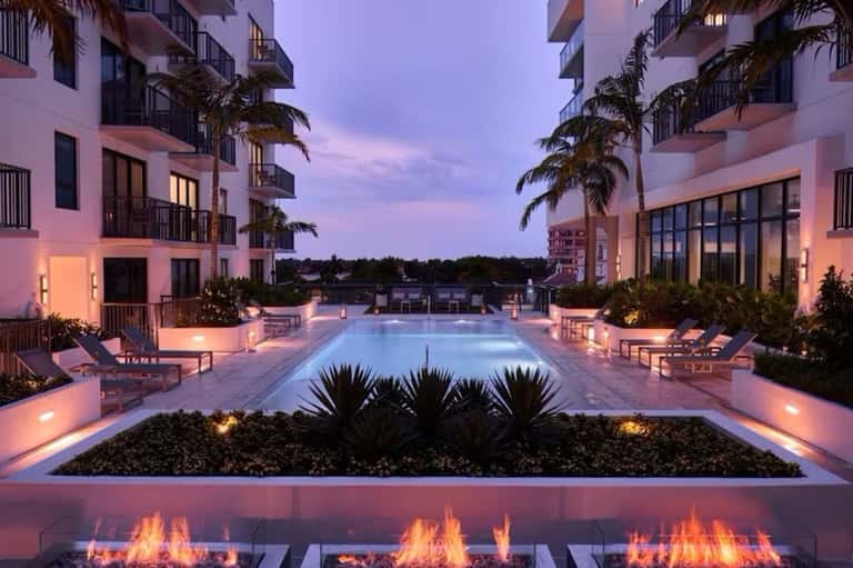 Illuminated resort pool at dusk surrounded by pink buildings, lounge chairs, and fire features