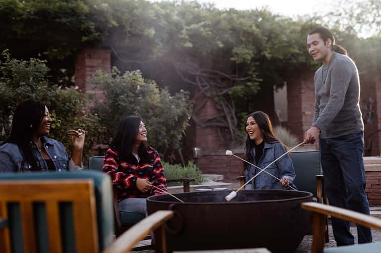 Group of friends gathering around a fire pit on a brick patio with autumn foliage in background