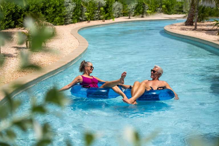 Couple floating on blue tubes in a lazy river pool surrounded by sandy banks and palm trees