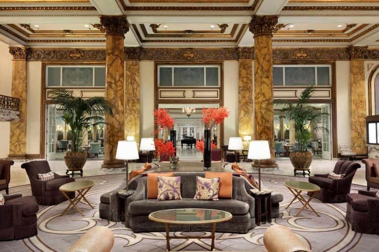 Luxurious hotel lobby with ornate columns, high ceilings, red floral arrangements, and elegant seating