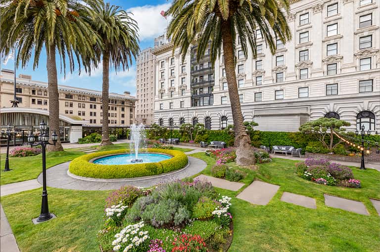 Manicured courtyard with fountain, palm trees, flowering plants, and classical buildings in background